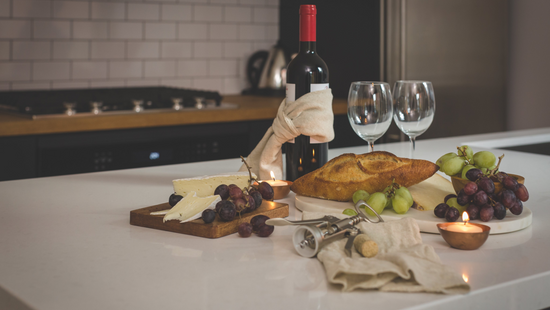 Wine bottle, glasses, bread, and fruits on a kitchen counter with a candle. 